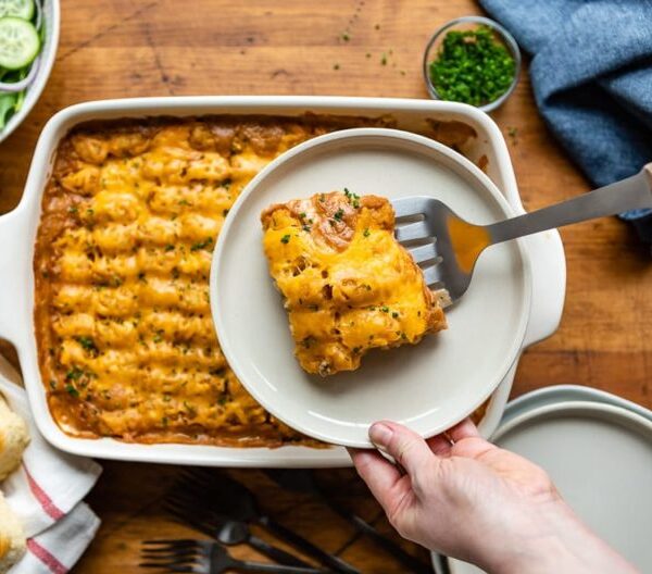 A hand holds a plate with a serving of cheesy casserole above a baking dish filled with more casserole, set on a wooden table. Various side dishes surround the baking dish.