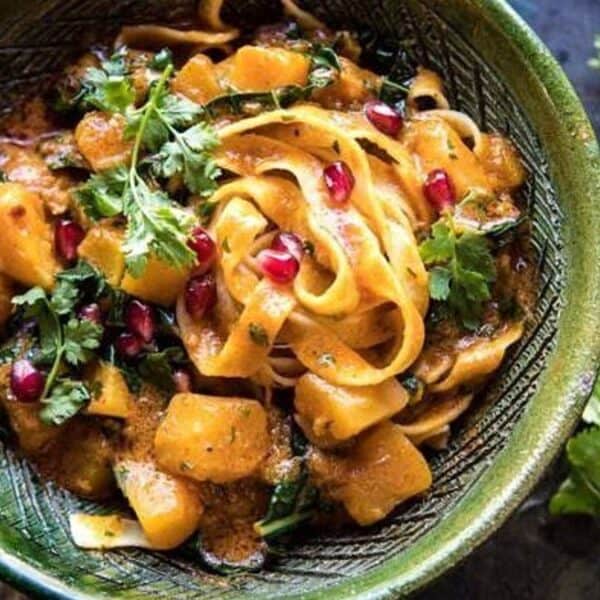 Green bowl holding noodles, squash chunks, fresh cilantro, and pomegranate seeds, with chopsticks and loose cilantro on a dark surface.