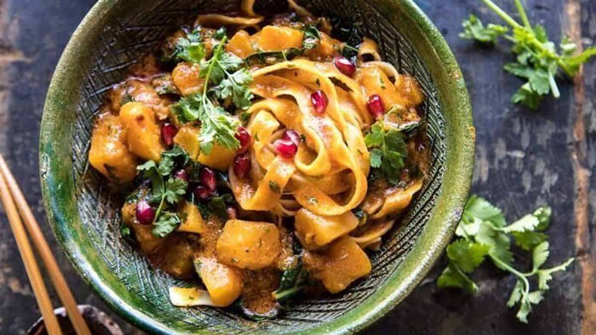 Green bowl holding noodles, squash chunks, fresh cilantro, and pomegranate seeds, with chopsticks and loose cilantro on a dark surface.
