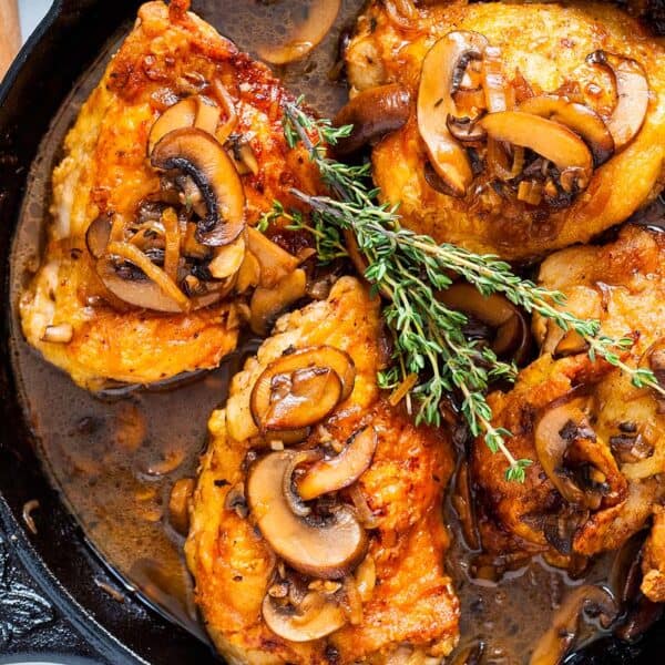 Four browned chicken thighs, mushrooms, and fresh thyme in a cast iron skillet.