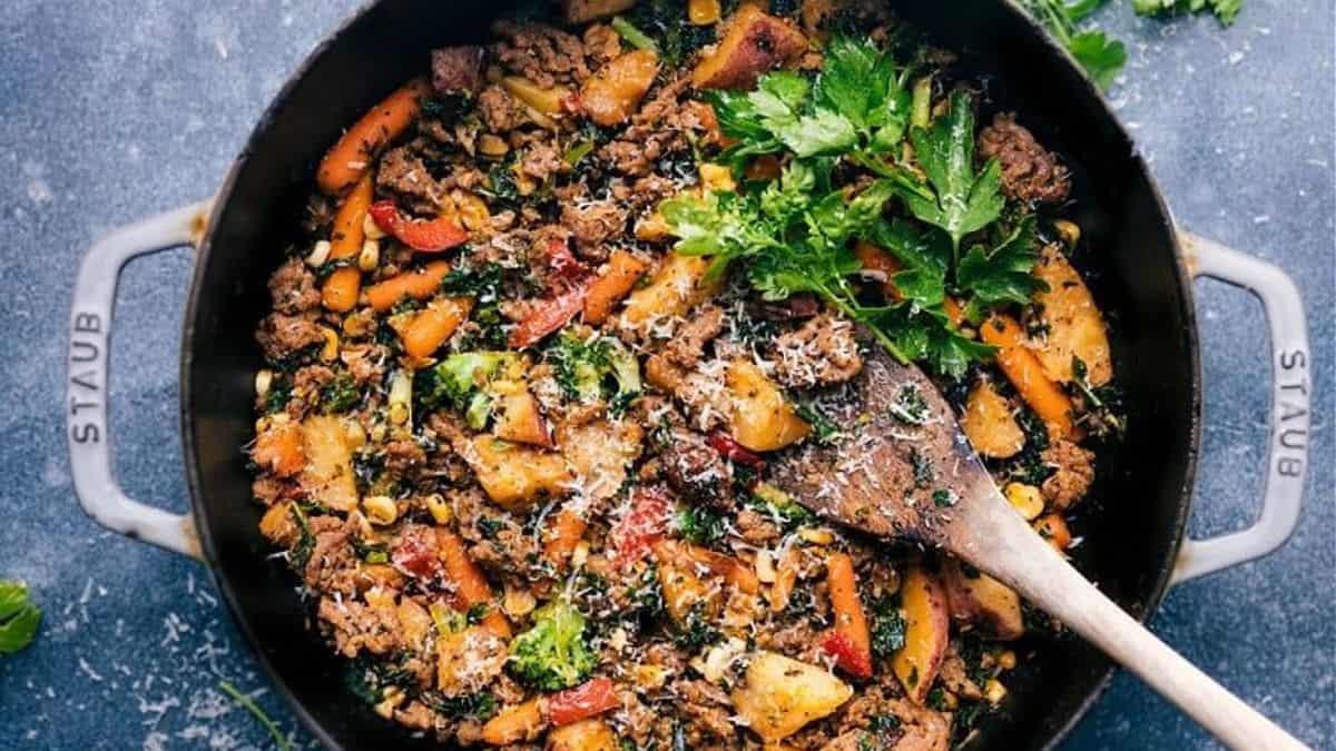 A large black Staub pot containing ground meat, vegetables, grated cheese, fresh parsley, and a wooden spoon inside.