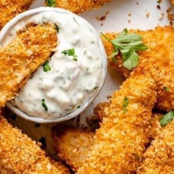Breaded baked chicken strips on a plate with parsley garnish and a bowl of creamy white dipping sauce, one strip being dipped.