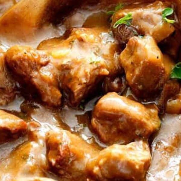 Chunks of beef in brown gravy, topped with chopped parsley, with part of a wooden spoon showing.