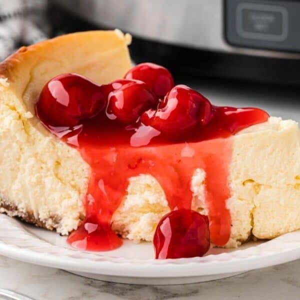 A slice of cheesecake with cherry sauce on a white plate, a fork next to it, and a checkered cloth in the background.