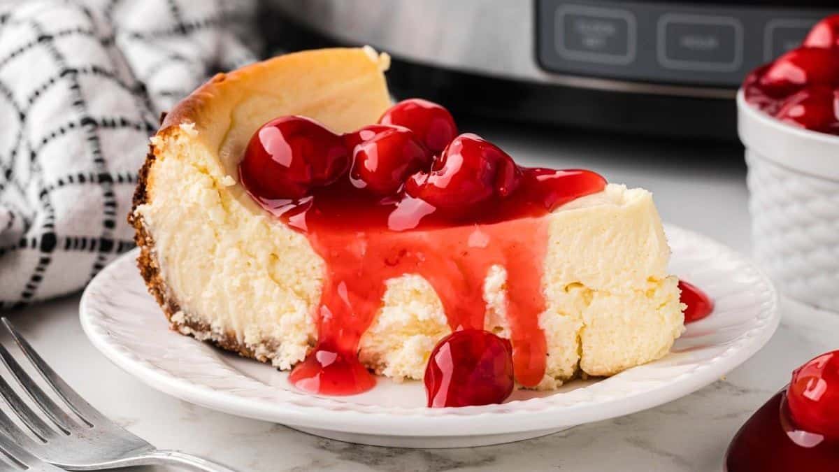 A slice of cheesecake topped with cherry sauce and whole cherries sits on a white plate, with a fork and a checkered cloth nearby.
