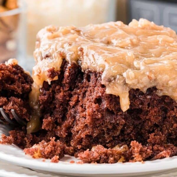 A close-up of a slice of chocolate cake with thick caramel and nut topping on a white plate, a fork breaking off a piece.