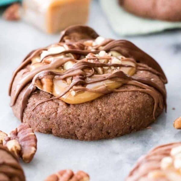 Chocolate cookie with caramel, chopped pecans, and chocolate drizzle, surrounded by pecan pieces on a light-colored surface.