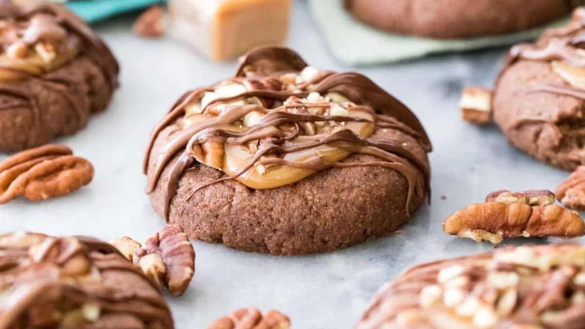 Chocolate cookie with caramel, chopped pecans, and chocolate drizzle, surrounded by pecan pieces on a light-colored surface.