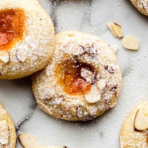 Jam-filled thumbprint cookies topped with powdered sugar and almond slices are arranged on a white surface.