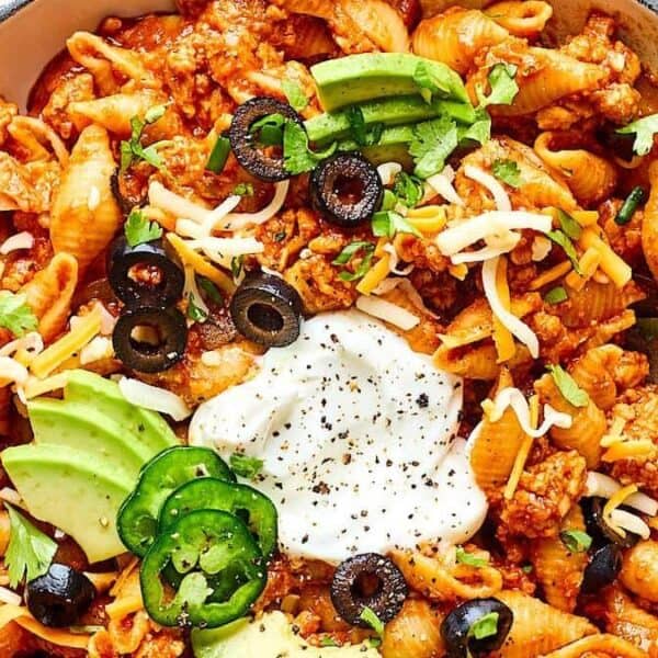 Taco pasta shells in a skillet topped with avocado slices, black olives, shredded cheese, sour cream, jalapeños, and cilantro.