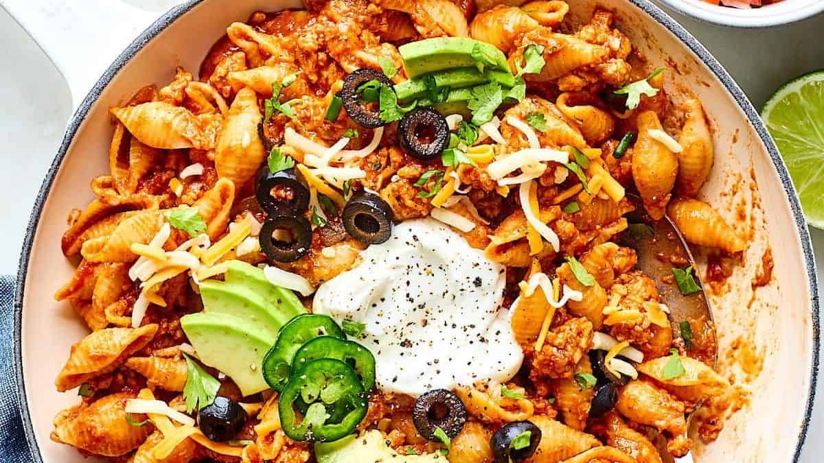 Taco pasta shells in a skillet topped with avocado slices, black olives, shredded cheese, sour cream, jalapeños, and cilantro.