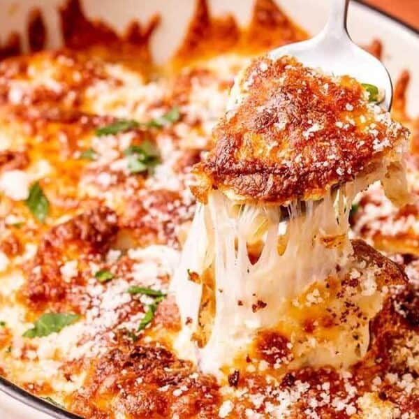A spoon holds a slice of cheesy baked lasagna above a casserole dish, with stretchy melted cheese and herbs visible on top.