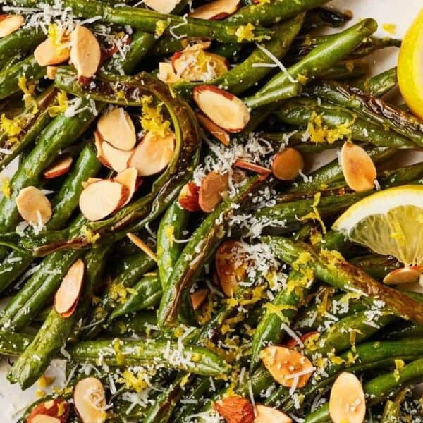 A plate of roasted green beans topped with sliced almonds, grated cheese, lemon zest, and lemon wedges on the side.