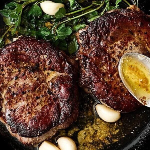 Two seared steaks in a cast iron pan with garlic cloves, fresh herbs, and melted butter.