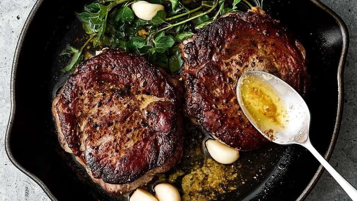Two seared steaks in a cast iron pan with garlic cloves, fresh herbs, and melted butter.