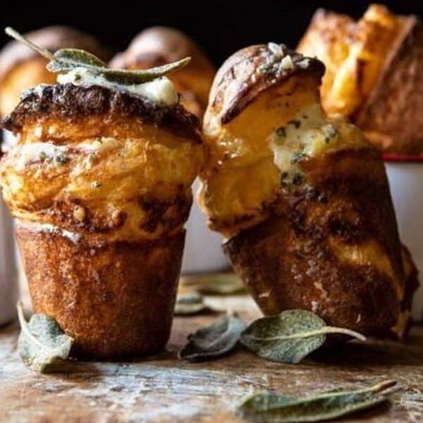 Golden popovers in white ramekins on a wooden surface, with melted butter and fresh sage leaves on top.