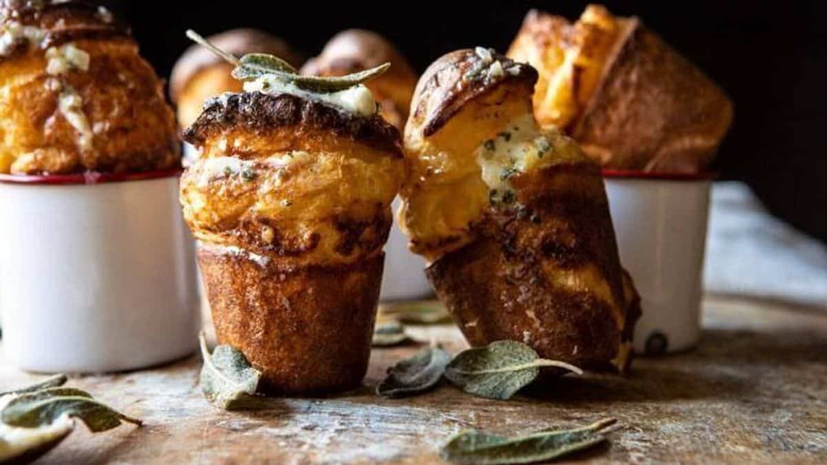Golden popovers in white ramekins on a wooden surface, with melted butter and fresh sage leaves on top.