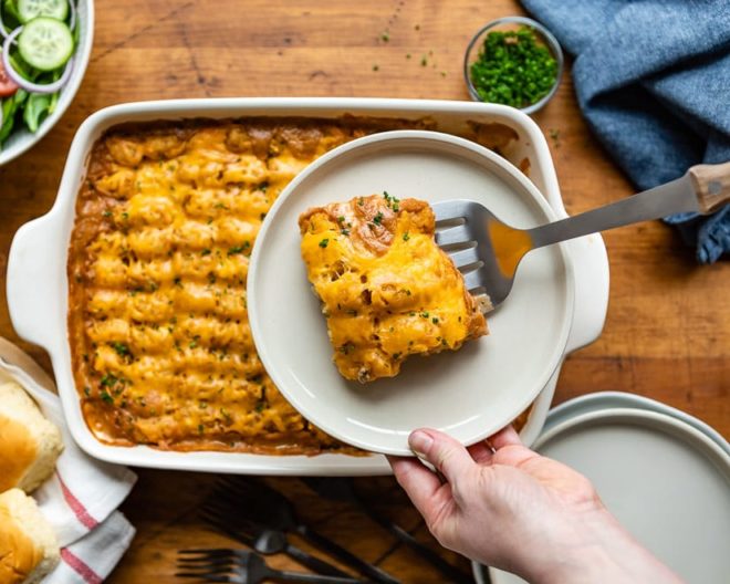 A hand holds a plate with a serving of cheesy casserole above a baking dish filled with more casserole, set on a wooden table. Various side dishes surround the baking dish.