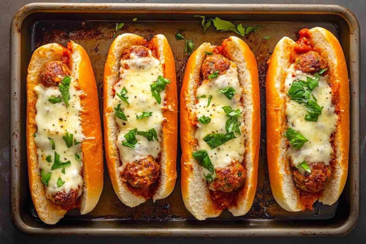 Four toasted sub rolls filled with meatballs, marinara sauce, and melted cheese, topped with chopped parsley, arranged on a baking sheet.