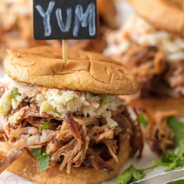 A pulled pork sandwich with coleslaw on a bun, topped with a small sign that says "YUM," rests on leafy greens on a wooden surface.