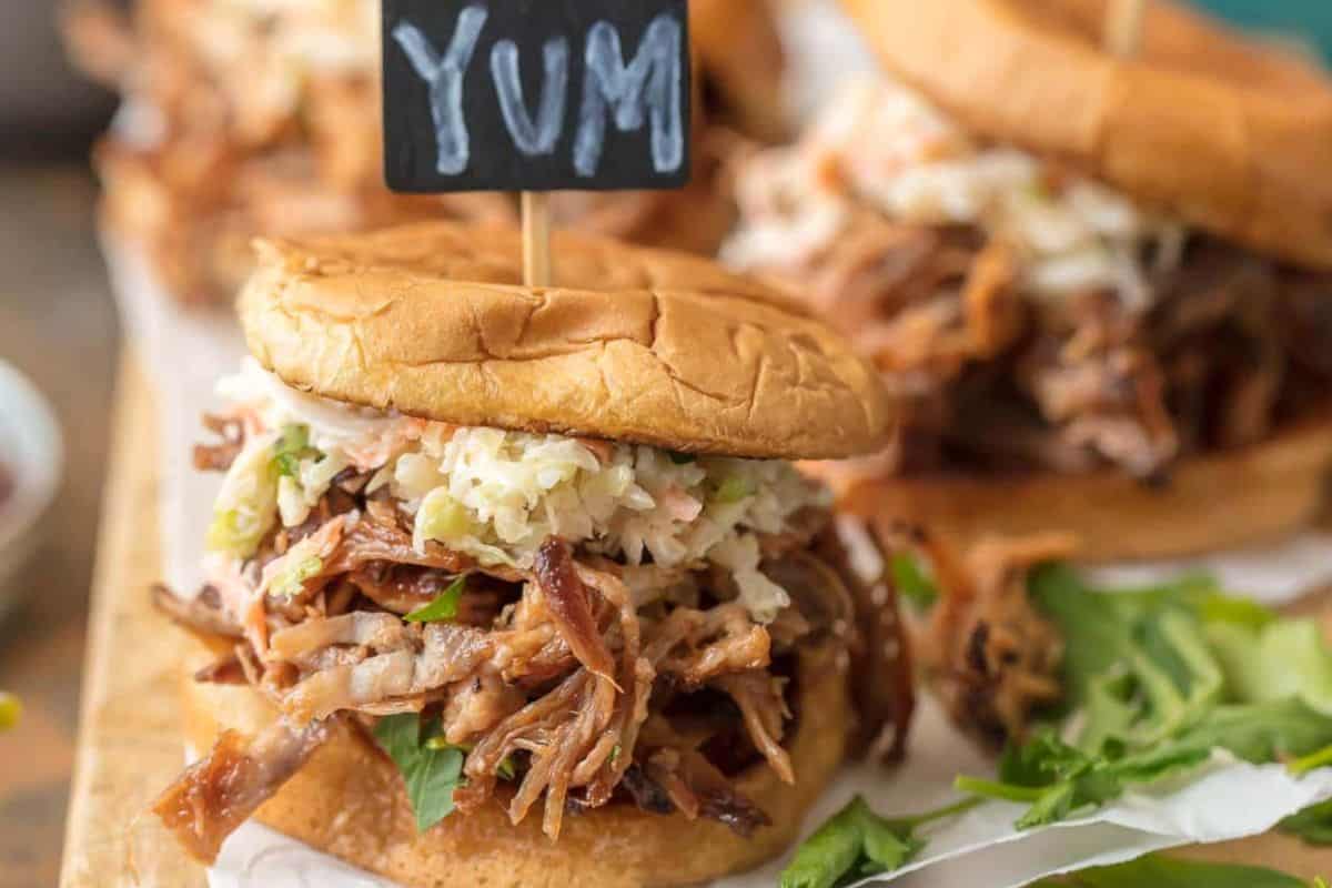 A pulled pork sandwich with coleslaw on a bun, topped with a small sign that says "YUM," rests on leafy greens on a wooden surface.