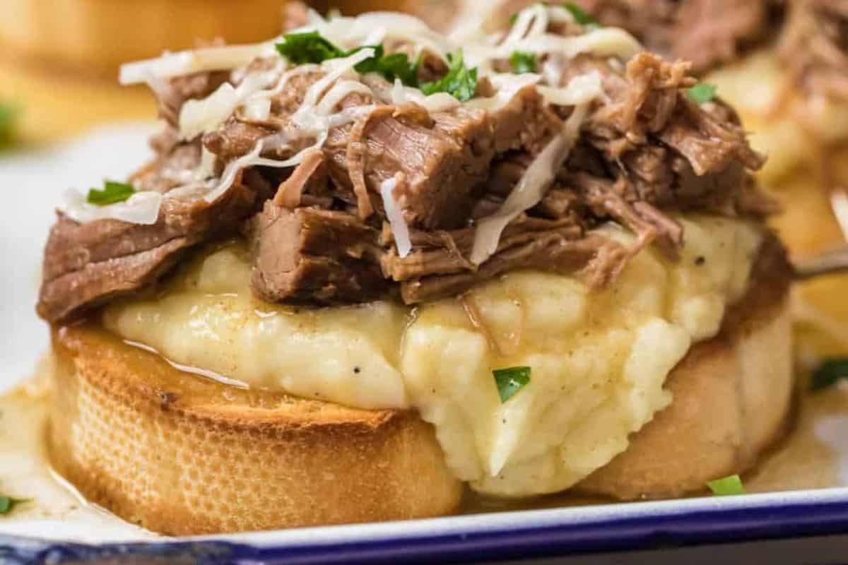 A slice of toasted bread topped with mashed potatoes, shredded beef, grated cheese, and fresh herbs on a white plate.