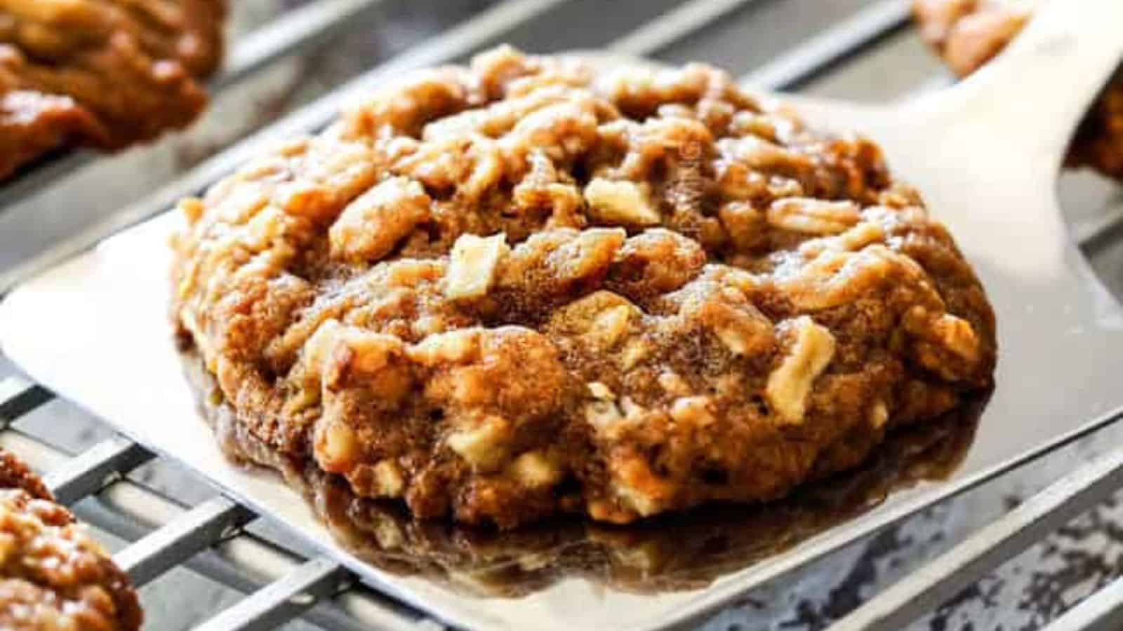 A homemade oatmeal cookie with oats on a metal spatula above a wire cooling rack.
