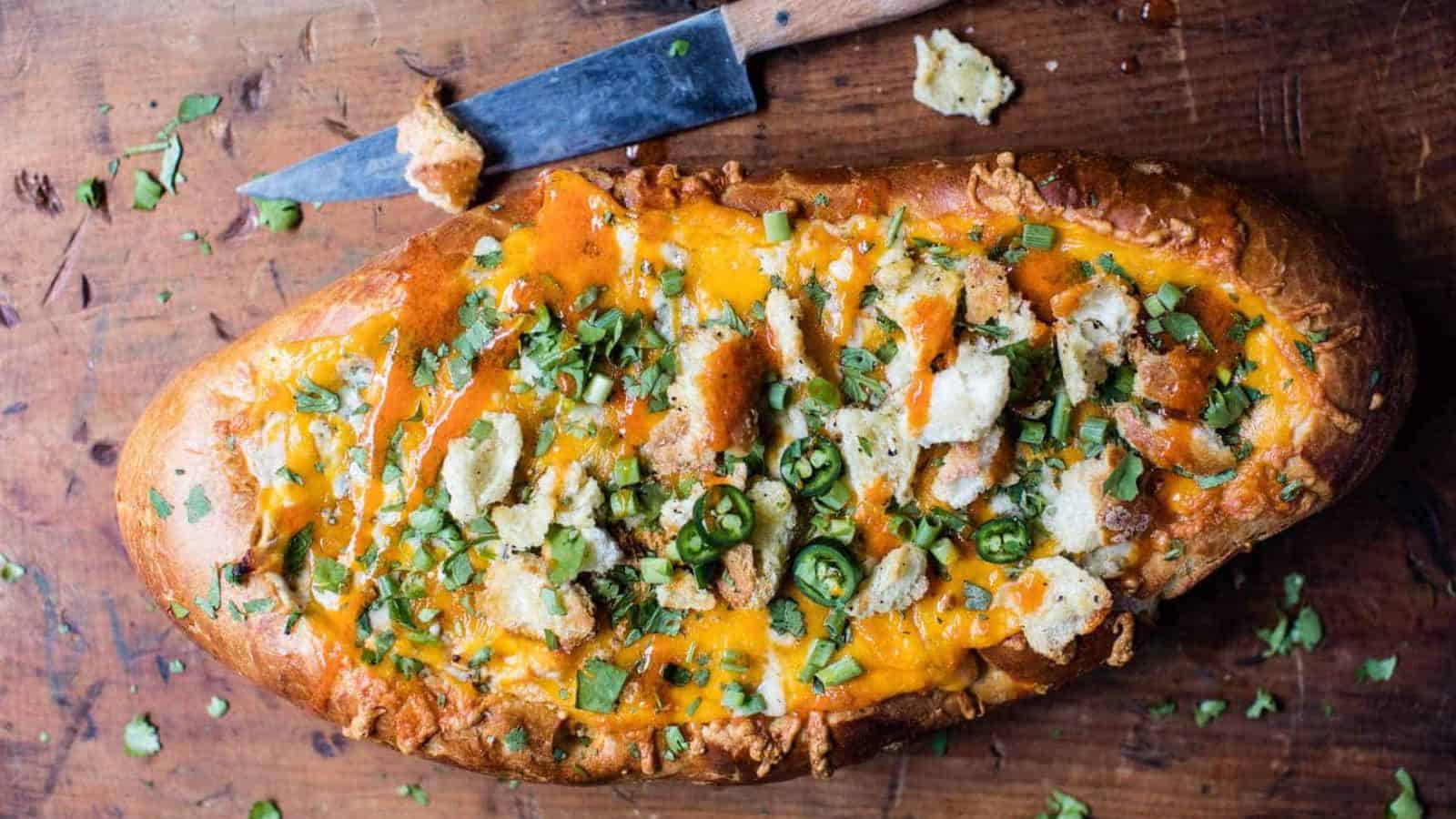 Baked bread loaf with melted cheese, green herbs, sliced jalapeños, crumbles, on wood surface beside a knife.