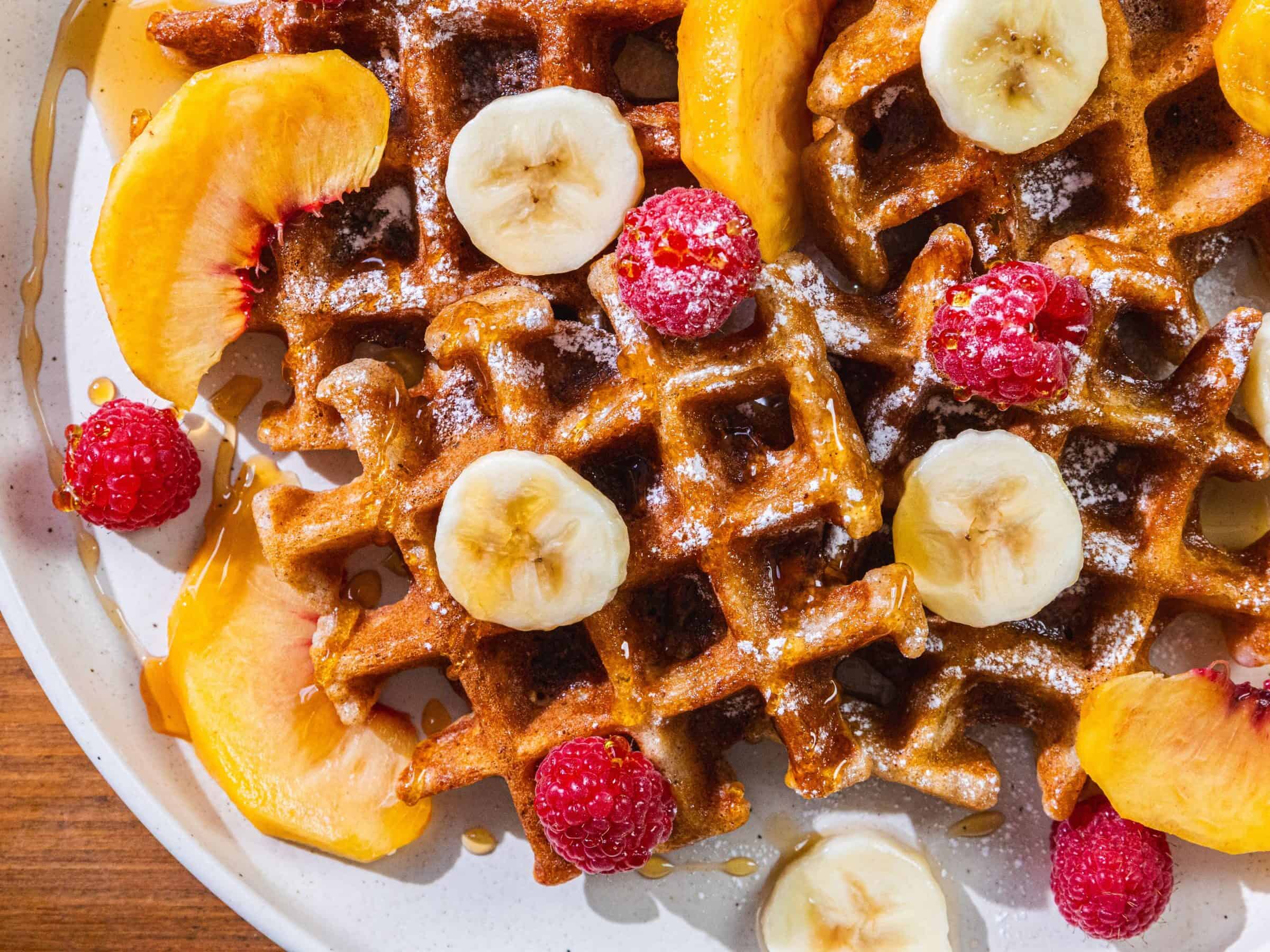 Waffles with banana slices, raspberries, peach slices, powdered sugar, and syrup on a white plate.