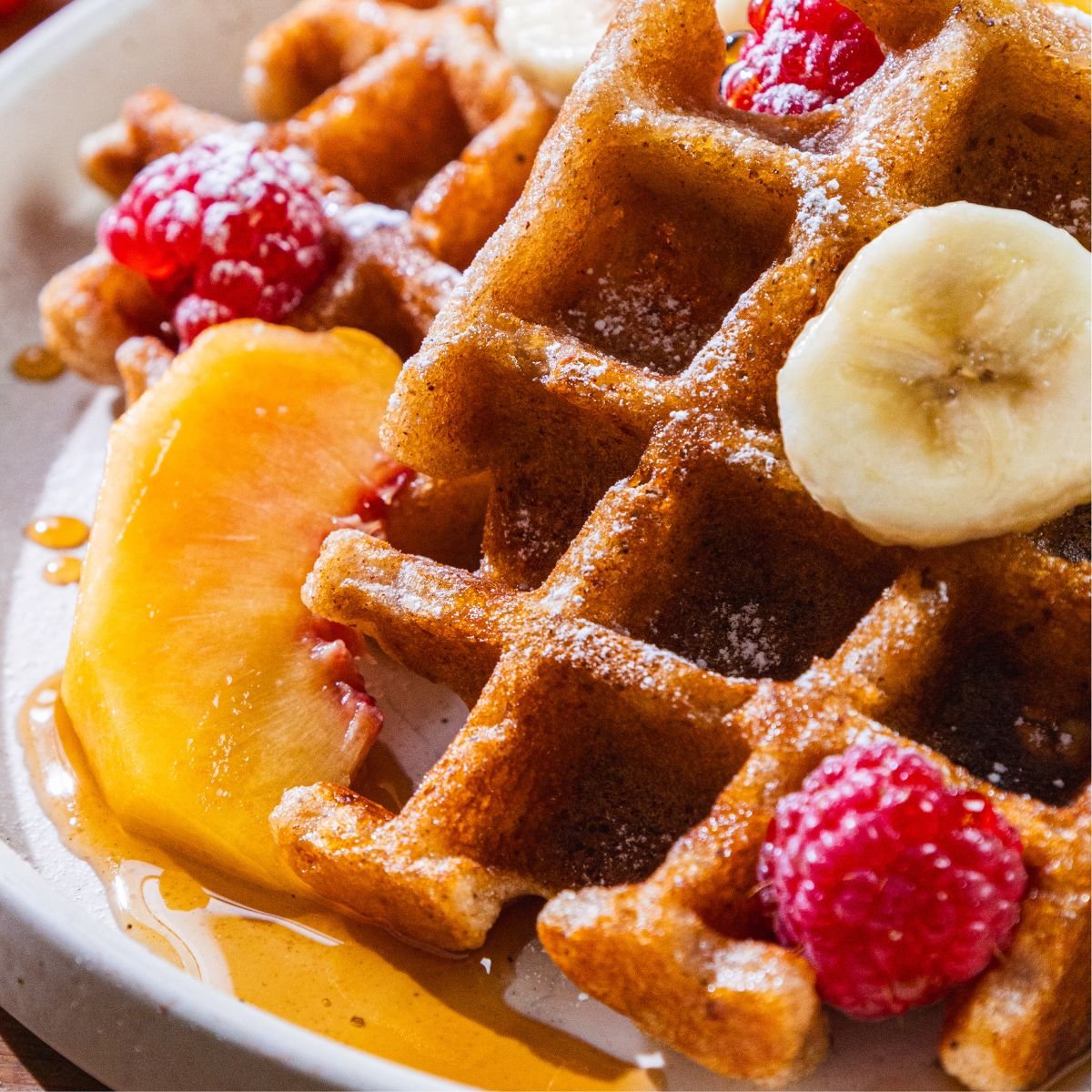 Waffles on a plate with banana slices, raspberries, a peach slice, and syrup.