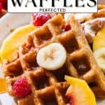 Plate of waffles with banana slices, peach slices, raspberries, and powdered sugar on top.