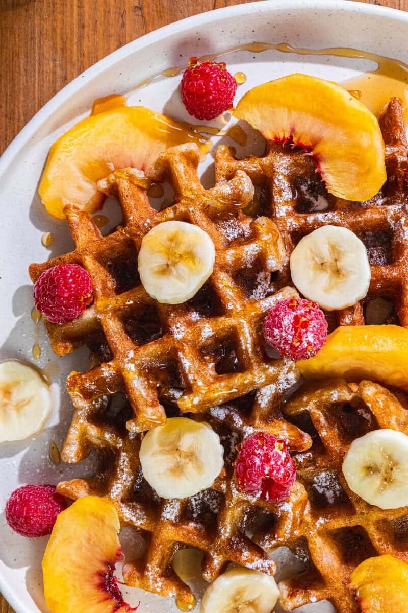 Waffles with banana slices, raspberries, peach slices, and syrup on a white plate.
