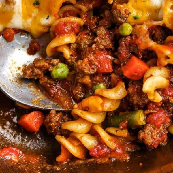 Cooked rotini pasta, ground beef, tomato sauce, peas, carrots, corn, and melted cheese in a casserole dish with a serving spoon.