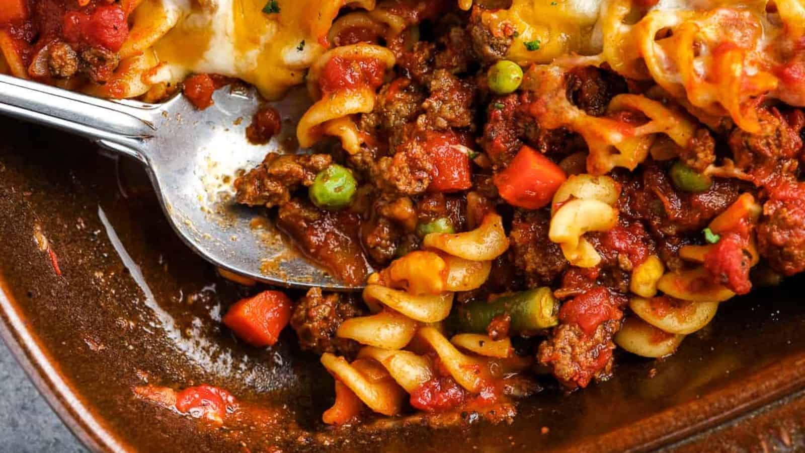 Cooked rotini pasta, ground beef, tomato sauce, peas, carrots, corn, and melted cheese in a casserole dish with a serving spoon.