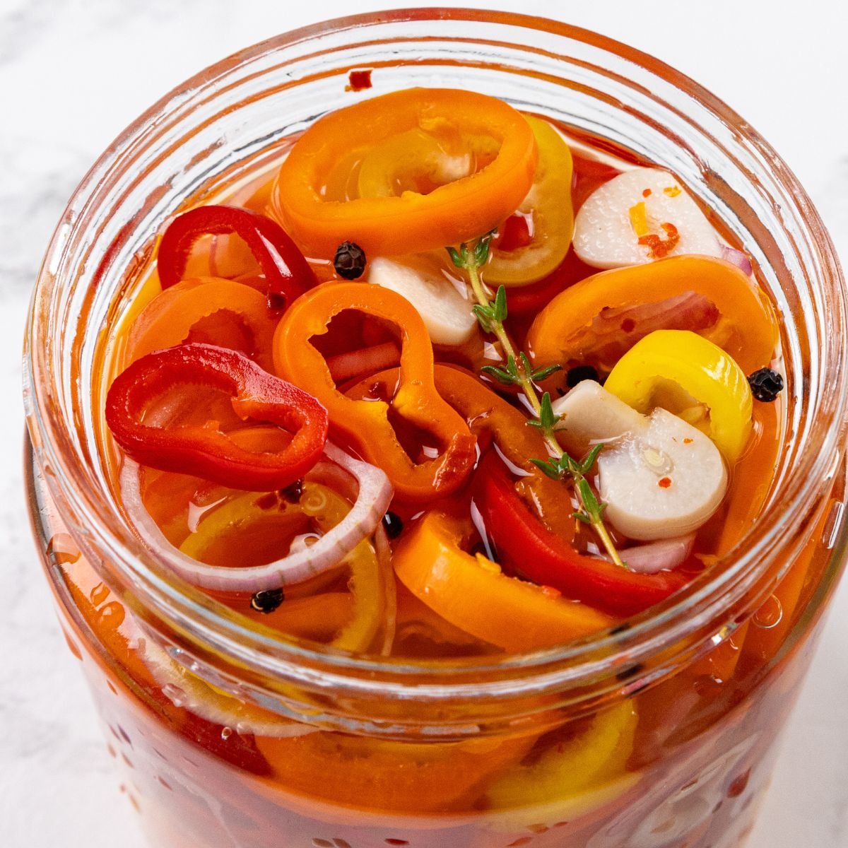 A glass jar containing sliced red, orange, and yellow peppers, garlic cloves, peppercorns, thyme, and pickling liquid.