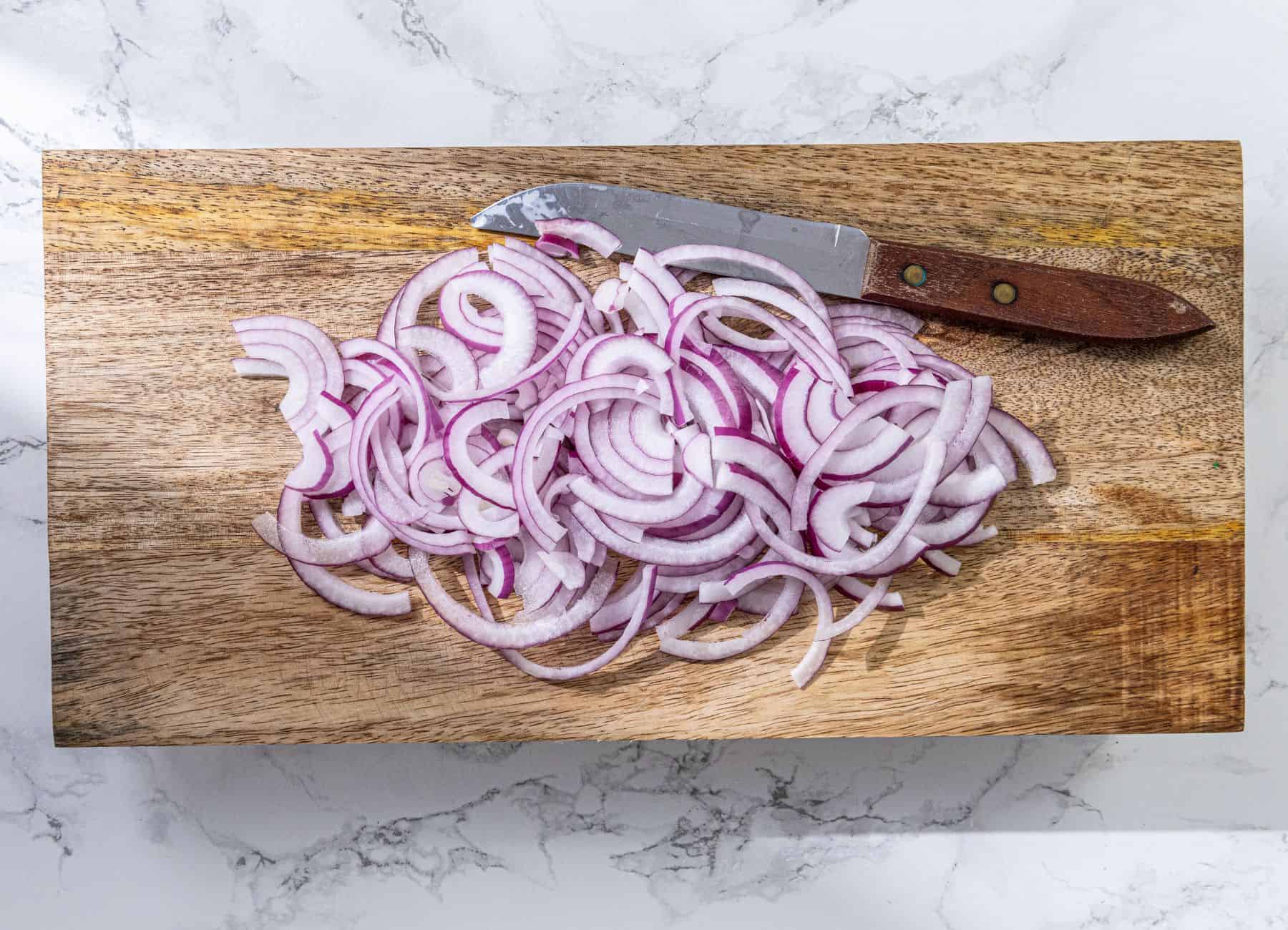 Sliced red onions and a knife rest on a wooden cutting board atop a white marble surface.
