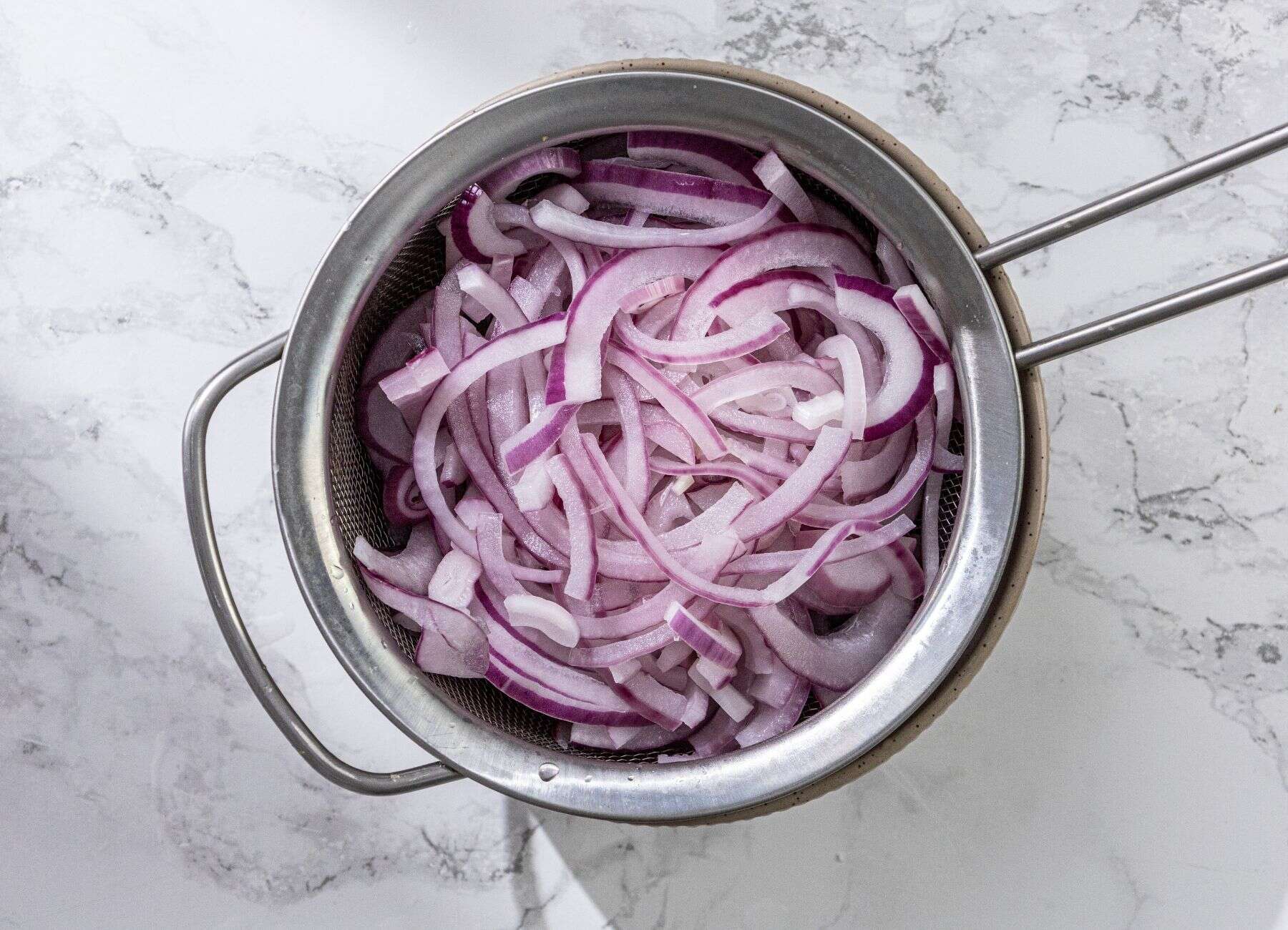 Sliced red onions in a metal strainer on a white marble surface.