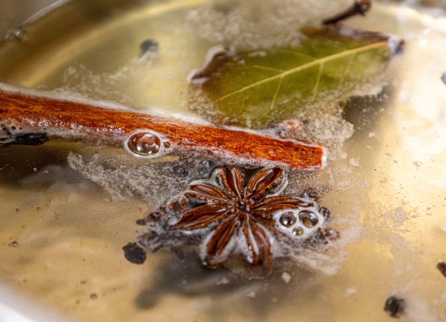 Cinnamon stick, bay leaf, star anise, and peppercorns floating on the surface of a simmering liquid.