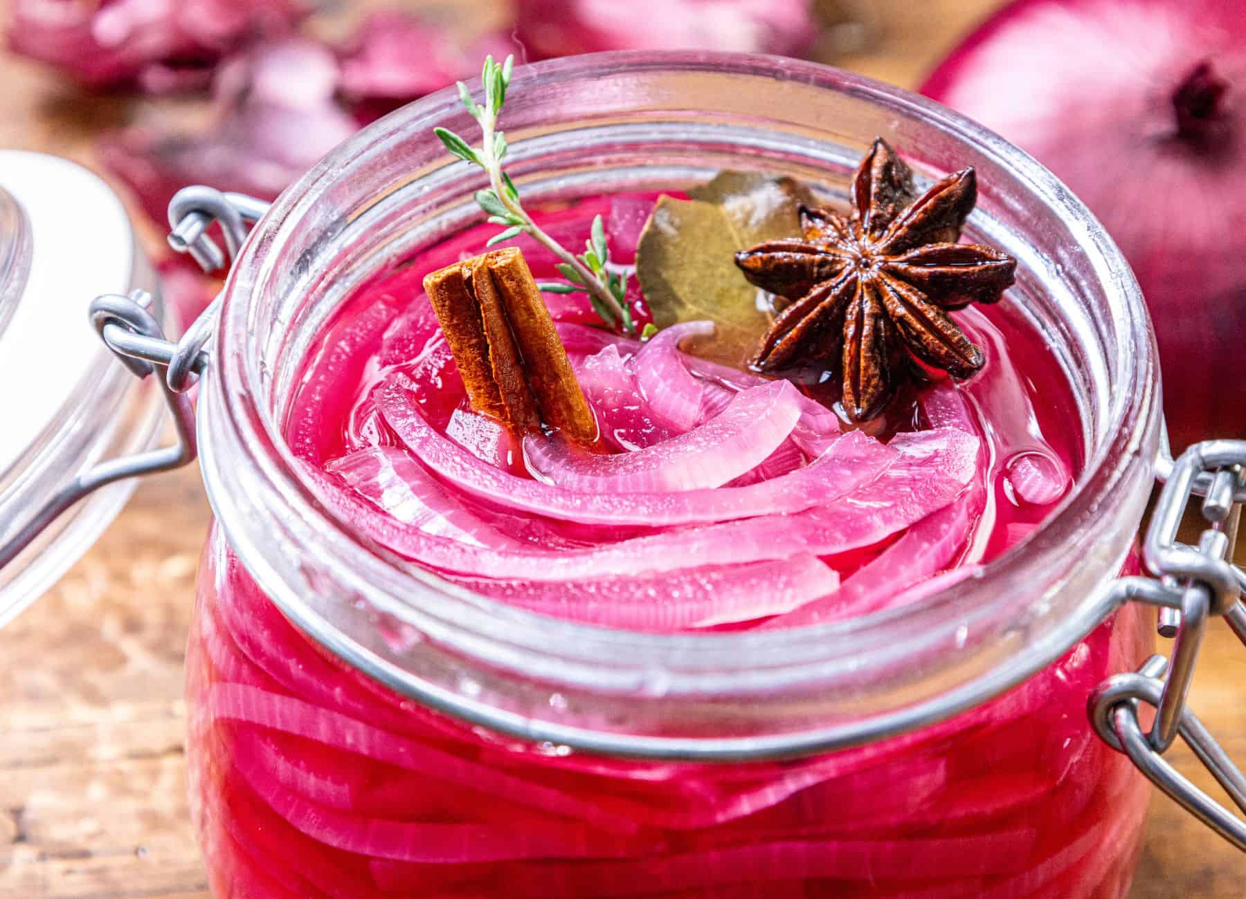 A glass jar of pickled red onions with cinnamon stick, star anise, bay leaf, and thyme on a wooden surface.