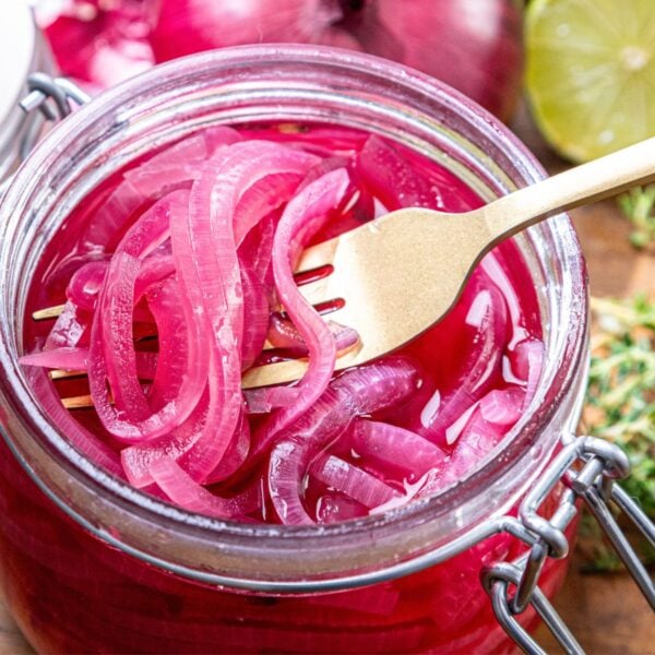 A jar of sliced pickled red onions with a fork in it, fresh lime and herbs arranged around the jar.