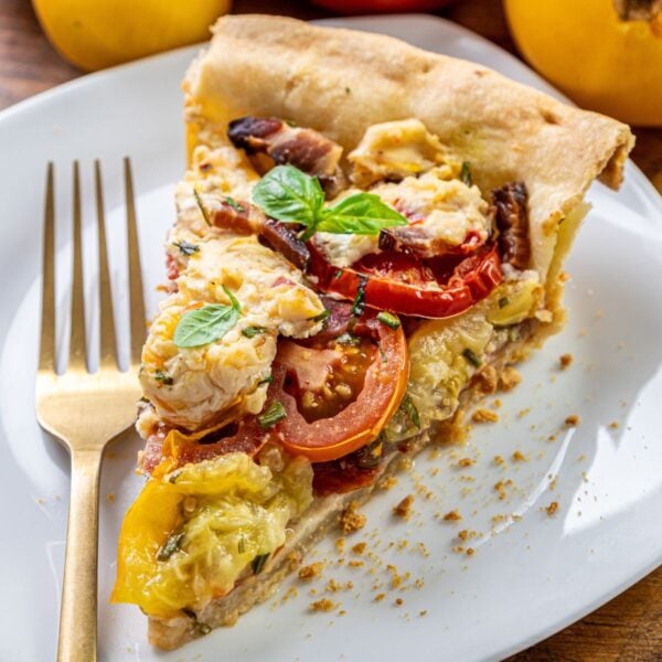 A slice of tomato and cheese pie with basil on top sits on a white plate next to a gold fork.