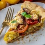 A slice of tomato and cheese pie with basil on a white plate, gold fork beside it, surrounded by whole tomatoes and basil leaves.
