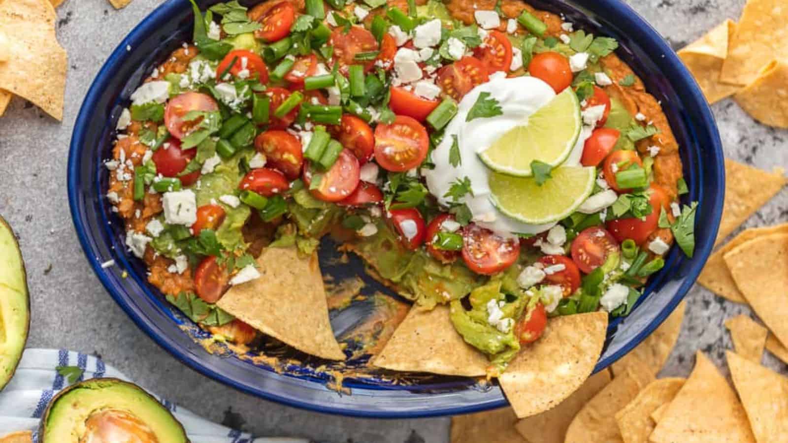 Blue dish filled with layered dip, topped with guacamole, cherry tomatoes, green onions, sour cream, lime slices, and tortilla chips.