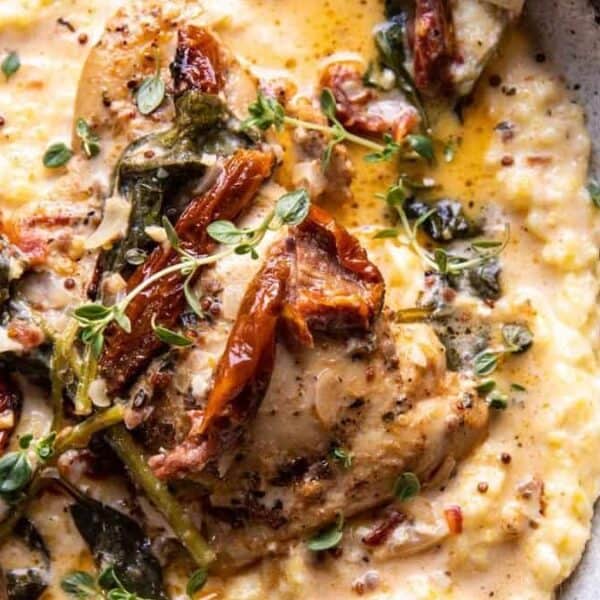 A bowl of creamy polenta with cooked chicken, sundried tomatoes, herbs, creamy sauce, and a fork on the side.