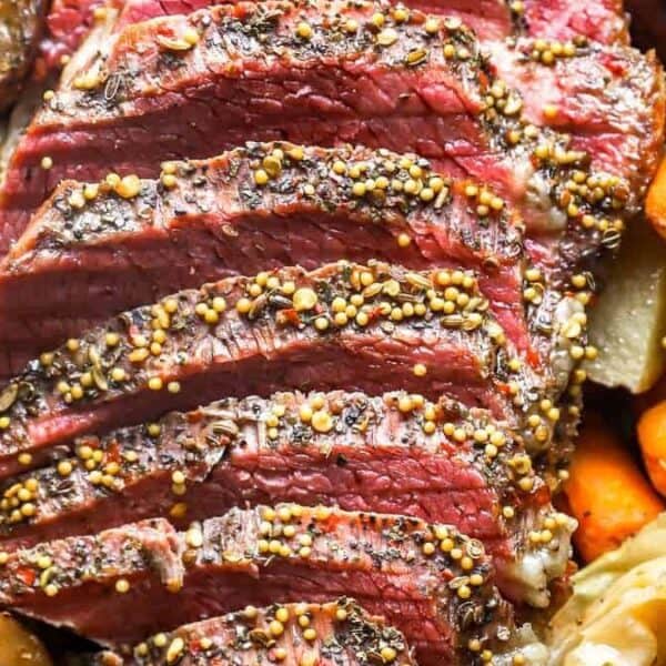 Sliced corned beef topped with mustard seeds, surrounded by cooked carrots, potatoes, cabbage, and onions.