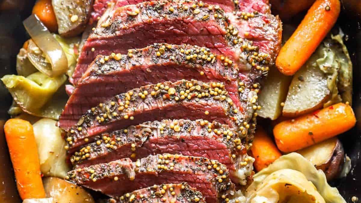 Sliced corned beef topped with mustard seeds, surrounded by cooked carrots, potatoes, cabbage, and onions.