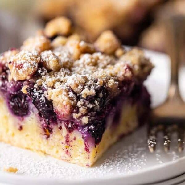 A slice of blueberry crumb cake with powdered sugar on a white plate, next to a fork.