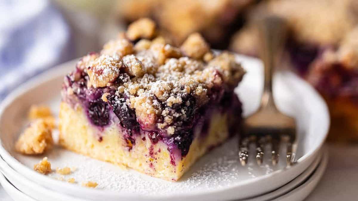 A slice of blueberry crumb cake with powdered sugar on top sits on a white plate, a fork placed next to it.