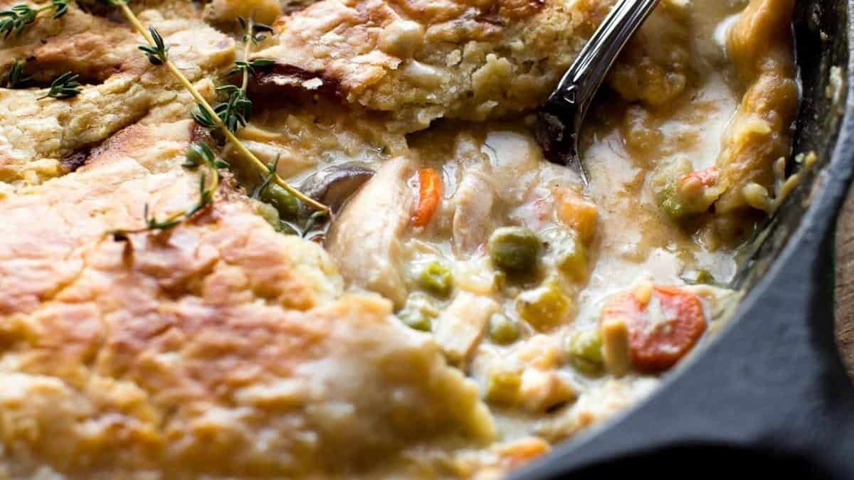 A skillet chicken pot pie with a golden flaky crust, creamy filling, carrots, peas, and a serving spoon.
