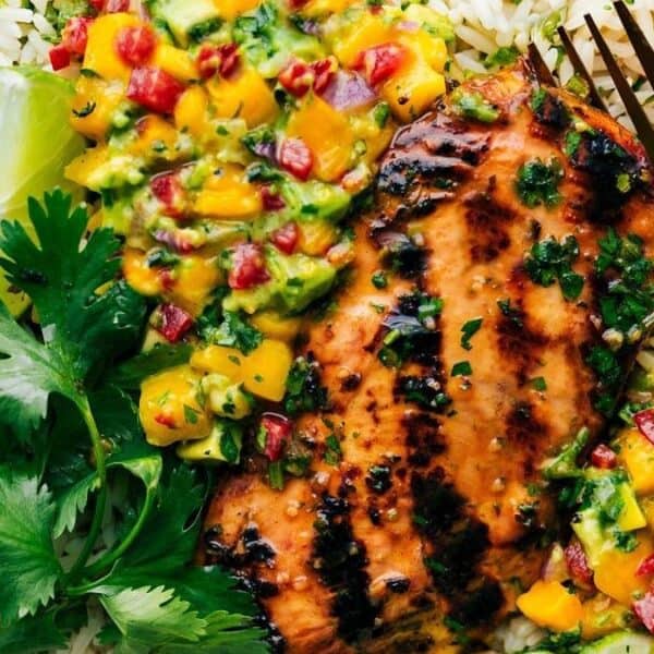 Grilled chicken breast on white rice with mango salsa, cilantro garnish, lime wedge, and a gold fork on the side.